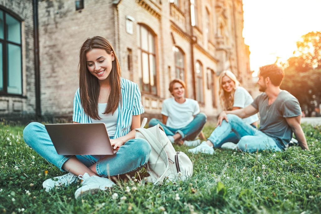 Studentenkonto - Studentin sitzt vor der Uni mit ihren Laptop auf dem Rasen